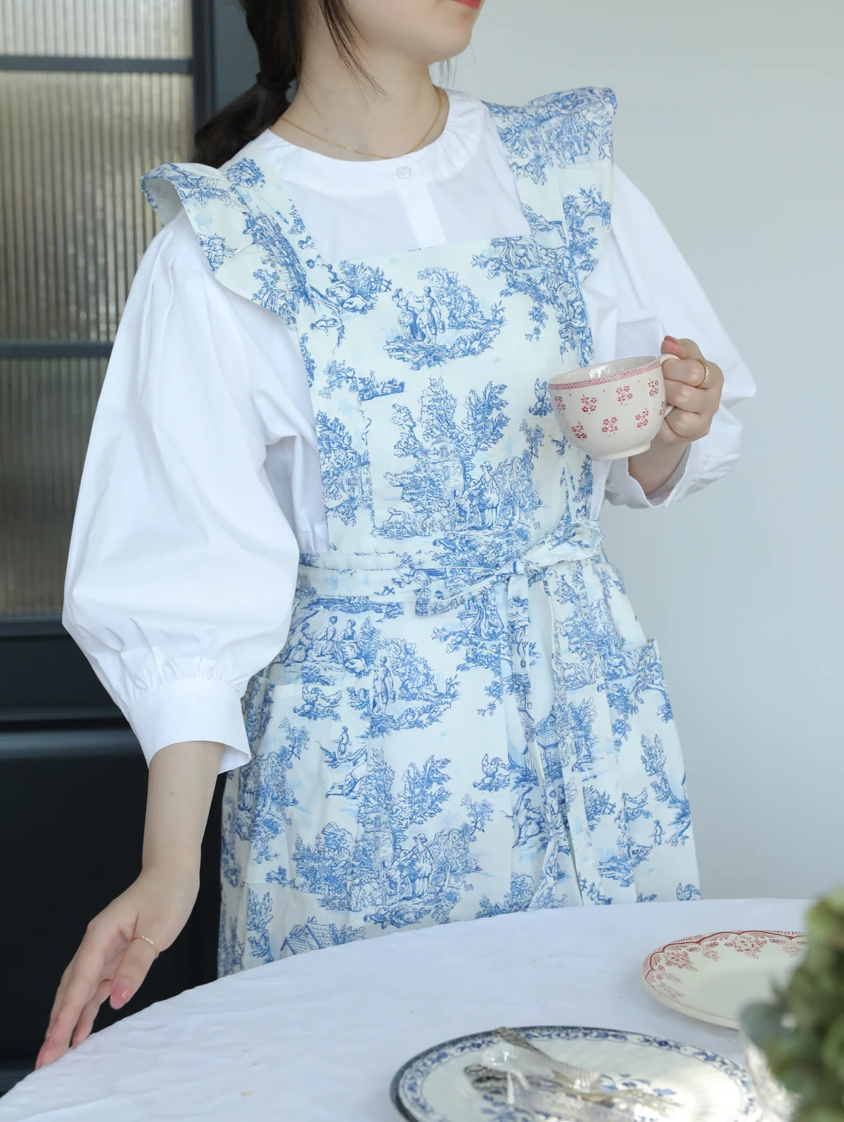 Wearing Hanee’s blue and white toile ruffle apron while holding a teacup beside a dining table